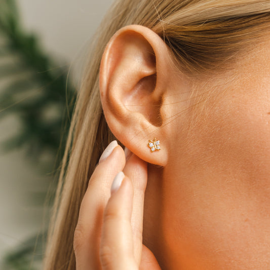 Lifestyle shot of the BLOOMTINE gold butterfly moissanite studs on a woman's ear. These gold-vermiel-over-silver earrings showcase the perfect scale for a dainty yet impactful look, highlighting the tarnish-resistant finish and high-brilliance stones against a soft-focus background