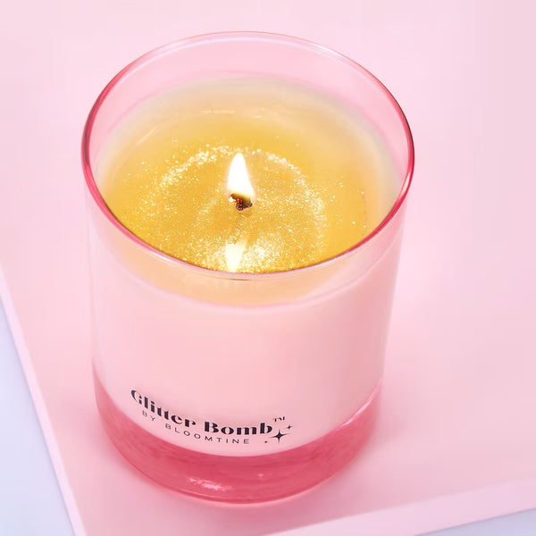 Pink candle labeled 'Glitter Bomb by Bloomtime' on a light pink background. The BLOOMTINE Glitter Bomb™ Gift Candle in a signature pink glass jar. This natural soy wax candle reveals a mesmerizing iridescent sparkle melt pool as it burns, releasing an addictive scent of creamy coconut, zesty lime, and vanilla sugar