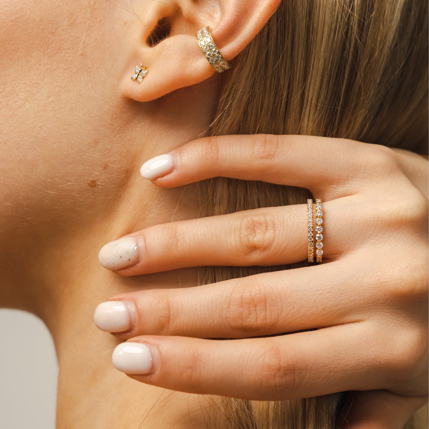 Close-up of a woman's hand and ear with gold jewelry including earrings and rings. The BLOOMTINE Illuminess™ Combi Ring in White Rhodium-plated gold vermeil. This exclusive two-in-one ring fuses a pavé band and a bezel-set moissanite into a single cohesive stack, offering a high-impact diamond-alternative look with a mirror-like, tarnish-resistant finish