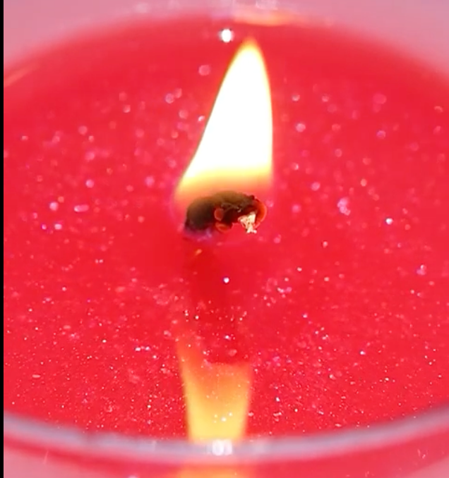 Rose-Galaxy-Glitter-Bomb-candle-close-up. A detailed-shot of a vibrant-red-scented-candle with a glowing-flame on the wick, surrounded-by-sparkling-gold-and-silver-glitter embedded-in-the-wax. The-image-features a warm-bokeh-background with soft-golden-lights, highlighting the-luxurious-shimmer-effect and cozy-atmosphere of this premium-Rose-and-Oud-gift-candle.